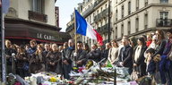 Menschen stehen vor dem Pariser Café Le Carillon, vor ihnen auf dem Boden haben Menschen Kerzen und Blumen abgelegt