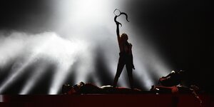 Die Silhouette von Madonna steht auf einer Bühne und hält einen gehörnten Kopfaufsatz in die Höhe, sie wird von unten angestrahlt