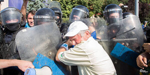 ein Mann mit weißer Baseballcap vor den Schutzschildern mehrerer Polizisten