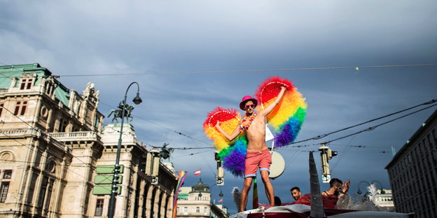 Ein Mann in Badehose steht auf einem Truck, er trägt einen pinken Hut und eine Blumenkette, an seinem Rücken prangen regenbogenfarbene Flügel