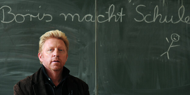 Boris Becker steht vor einer Schultafel mit der Aufschrift "Boris macht Schule".
