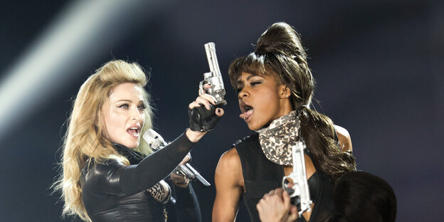Madonna steht auf einer Bühne, hält einen silbernen Revolver in der Hand, neben ihr eine Tänzerin