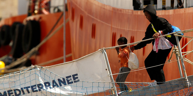 Eine Mutter im Kapuzenpulli mit einem kleinen Kind an der Hand geht von Bord eines Schiffes
