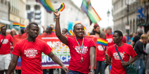 Drei Männer mit T-Shirts der „Rainbow Refugees Munich“, einer hält eine Regenbogenfahne