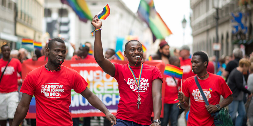 Drei Männer mit T-Shirts der „Rainbow Refugees Munich“, einer hält eine Regenbogenfahne