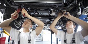 Zwei Männer in Arbeitskleidung stehen mit Werkzeug in der Hand unter einem Auto