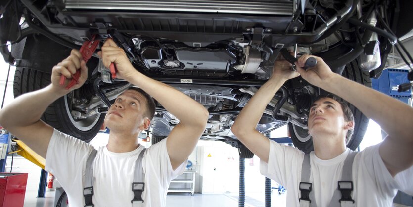 Zwei Männer in Arbeitskleidung stehen mit Werkzeug in der Hand unter einem Auto