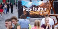 Menschen sitzen auf Bierbänken, im Hintergrund ein Plakat des Pop Kultur Festivals