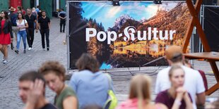 Menschen sitzen auf Bierbänken, im Hintergrund ein Plakat des Pop Kultur Festivals