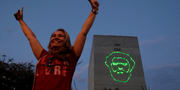 Eine Frau streckt erfreut die Arme in die Höhe, im Hintergrund ist das Gesicht von Lula mit grünen Lasern an eine Hauswand projeziert