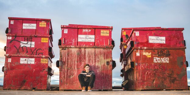 Ein Mensch hockt lächelnd vor drei roten, riesigen Müllcontainern