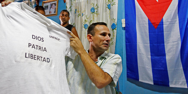 Der kubanische Dissident José Daniel Ferrer hält vor einer kubanischen Flagge ein T-Shirt mit der Aufschrift „Gott, Heimat, Freiheit“ in die Höhe