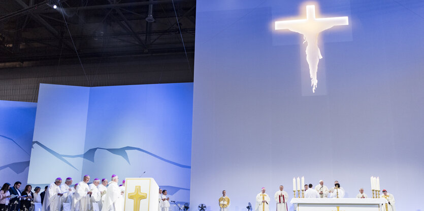 Katholische Würdenträger vor einer blau erleuchteten Wand mit einem strahlenden Riesenkreuz