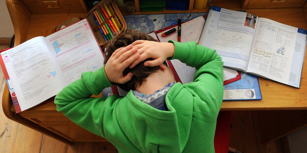 Ein Kind hat den Kopf zwischen seine Schulbücher auf den Tisch gelegt und die Hände im Nacken gefaltet.