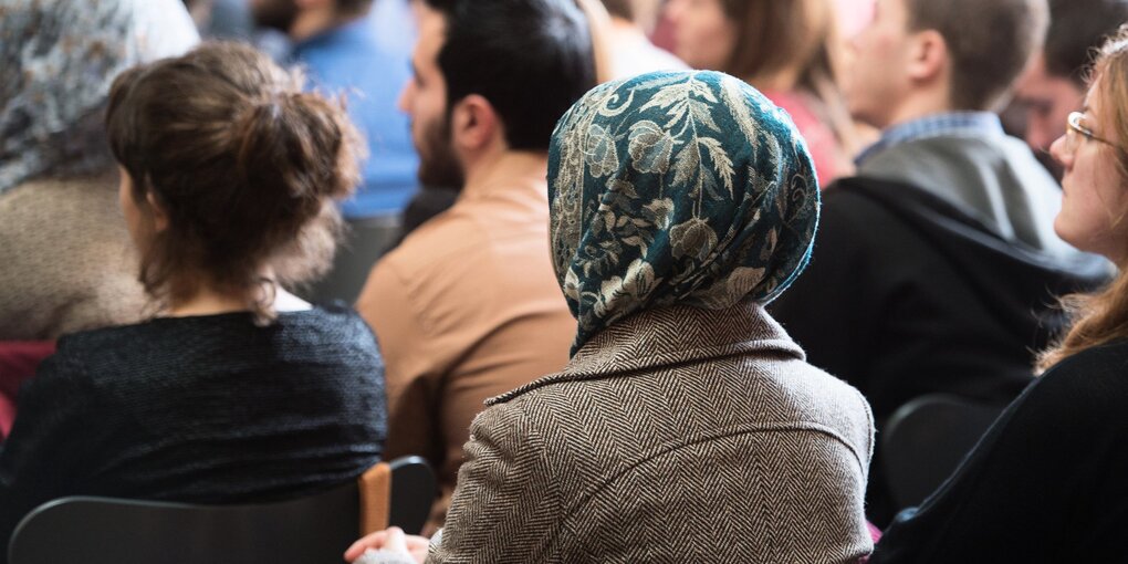 Eine Frau mit Kopftuch sitzt zwischen anderen Männern und Frauen, sie alle sitzen auf Stühlen, sind von hinten zu sehen und schauen geradeaus.