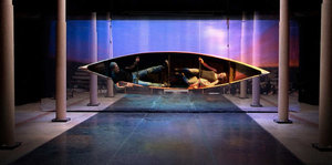 Auf einer Bühne hängt zwischen Säulen ein Kanu seitwärts, darin liegen ein Mann und eine Frau