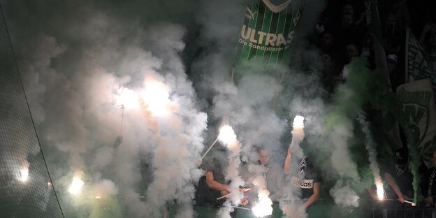Fans brennen Pyrotechnik ab und halten Fahnen hinter einer Nebelwand hoch