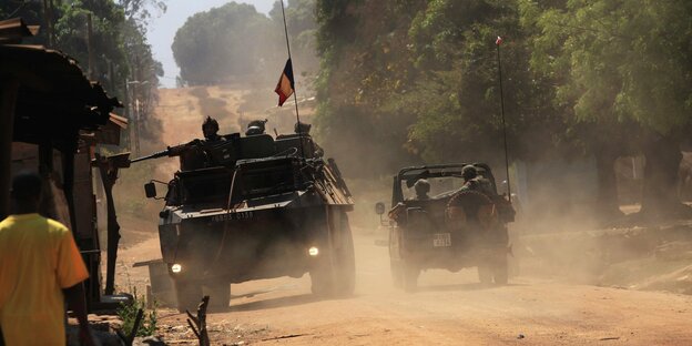 Ein Radpanzer mit französischer Flagge auf staubiger Straße in Bangui. Soldaten mit Schusswaffen darauf. Daneben ein Jeep. Links auf der Straße läuft ein Mann in gelbem Shirt.
