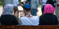 Zwei junge Muslimas mit Kopftuch und ein junger jüdischer Mann mit Kippa unterhalten sich miteinander