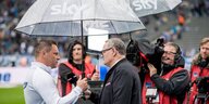 Zwei Männer auf einem Fußballplatz unter einem durchsichtigen Regenschirm. Der eine hält dem anderen ein Mikrofon vor das Gesicht