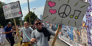 Junger Mann mit Sonnenbrille grinst und hält Schild hoch, darauf sind zwei Herzen, ein Peace-Zeichen und Noten zu sehen