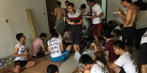 Eine Grupper junger Chinesen isst gemeinsam, teils auf dem Boden sitzend, manche tragen dasselbe Tshirt