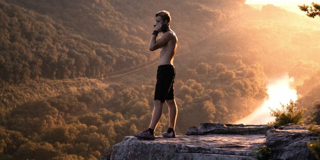 Ein Mann mit nacktem Oberkörper steht in goldenem Licht auf einer Klippe vor einer Berglandschaft
