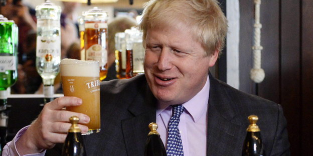 Boris Johnson hält ein Glas Bier in die Luft und grinst es an