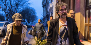 Yücel und seine Frau laufen über eine Straße, Männer im Hintergrund