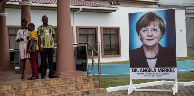 Ein PLakat, das Angela Merkel zeigt, steht neben einem Flughafengebäude. Neben dem Plakat stehen drei Menschen