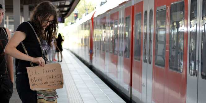 Eine Frau steht auf einem Bahnsteig. In ihrer Hand hält sie ein Schild, auf dem steht, dass sie schwarz fährt