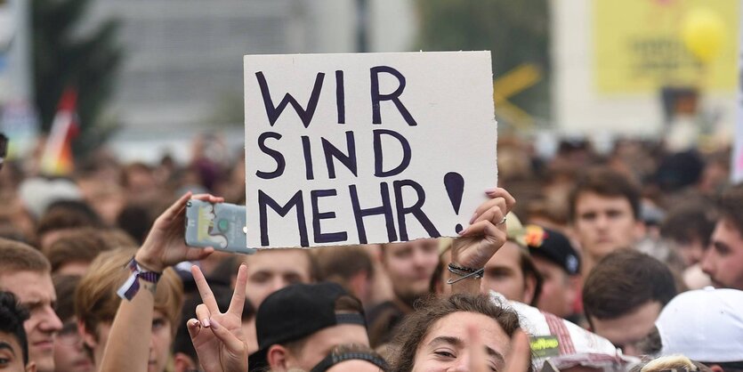 Köpfe von einer Menge Demonstranten. Aus der Menge heraus ragt ein Schild mit der Aufschrift „Wir sind mehr“