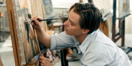 Tom Schilling mit einem Pinsel in der Hand vor einem gemalten Bild