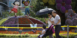 Vor einer Installation mit bunten Blumen sowie einer asiatischen und einer schwarzen Mädchenfigur läuft eine Familie mit Kinderwagen