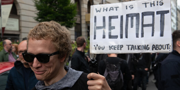 Ein Mann mit Sonnenbrille hält ein Plakat mit der Aufschrift „What is this Heimat you keep talking about“ hoch