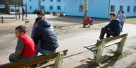 Kinder sitzen gelangweilt auf Bänken an einem Spielplatz vor kasernenartigen Gebäuden