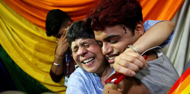 Vor einer Regenbogenflagge umarmen sich zwei Menschen, ihre Augen sind geschlossen, ihr Gesichtsausdruck erfreut