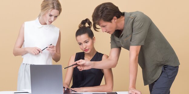 Eine Frau sitzt vor einem Laptop, rechts und links neben ihr stehen eine Frau und ein Mann, der auf den Bildschirm zeigt