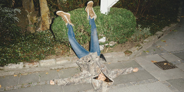 Gestelltes Foto einer Person, die auf dem Gehweg auf dem Rücken liegt, die Füße in einer Hecke