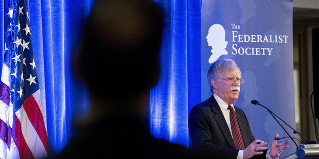 John Bolton steht an einem Rednerpult, vorne im Bild ist als dunkle Silhouette ein Mann von hinten zu sehen