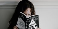 Ein Mädchen mit langen dunklen Haaren hat das Gesicht hinter einem Buch mit schwarzem Cover verborgen