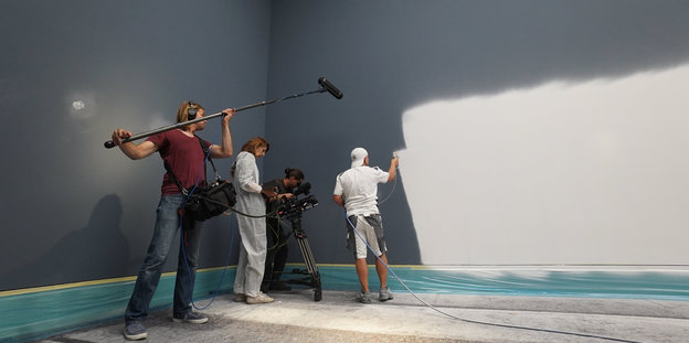 Ein Mann mit Baseballcap streicht eine graue Wand in einem großen Raum weiß an, dabei wird er von einem Kamerateam gefilmt