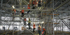 Elf Personen mit orangen Warnwesten steigen ein Baugerüst im Inneren der Neuen Nationalgalerie empor.