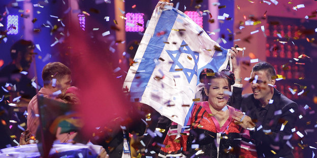 In einem Konfettiregen steht eine junge Frau, hinter ihr die israelische Flagge