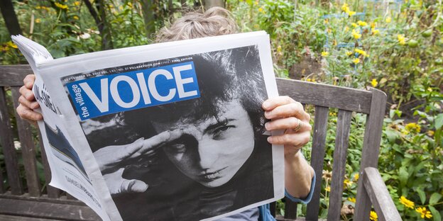 Eine Person sitzt auf einer Bank und hält eine Ausgabe der Zeitung "Village Voice" in der Hand