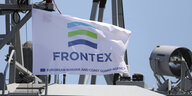 Eine weiße Flagge, auf der Frontex steht, im Hintergrund ein Schiff und blauer Himmel