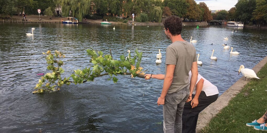 Zwei Männer stehen am Ufer eines Kanals, der eine hat einen großen Ast in der Hand, im Wasser schwimmen Schwäne