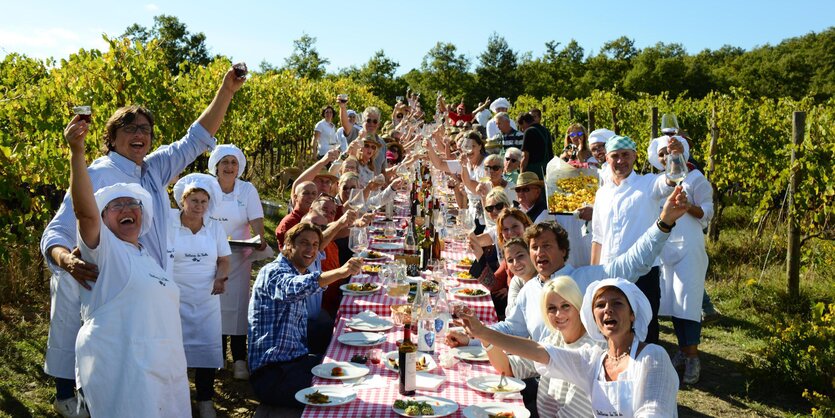 Mehrere Menschen stehen an einer langen Tafel in schöner Weinberg-Landschaft und prosten dem Betrachter zu