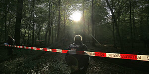 Ein Polizist steht vor einem rot-weißen Flatterband im Wald und guckt ins Sonnendurchflutete Dickicht