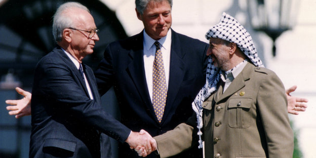 Arafat und Rabin schütteln sich die Hände, Bill Clinton breitet hinter ihnen die Arme aus.